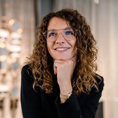 Kundin mit goldener Brille und Locken lächelt, Kinn auf die Hand gestützt, im Brillengeschäft